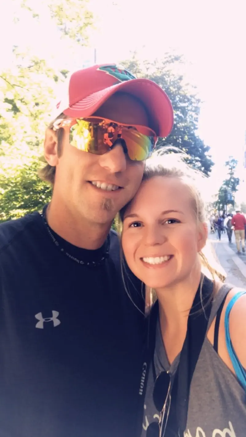 Smiling couple takes a sunny outdoor selfie; man in red cap and sunglasses, woman with blonde ponytail and camera strap, trees in background.