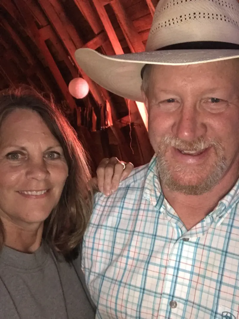 Smiling man in cowboy hat and plaid shirt stands indoors with a woman, who rests her hand on his shoulder under warm, reddish lighting.