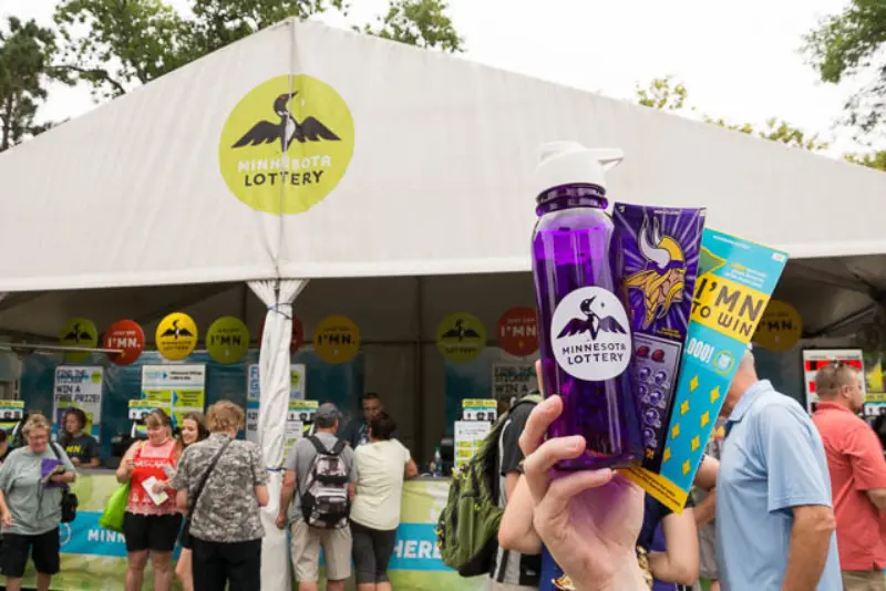Person displays a Minnesota Lottery water bottle, Vikings item, and lottery tickets at a busy outdoor Lottery booth with people under a white tent.