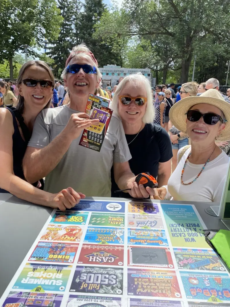 Four adults in sunglasses and a sunhat hold scratch-off tickets at an outdoor event with a lottery display board and trees in the background.