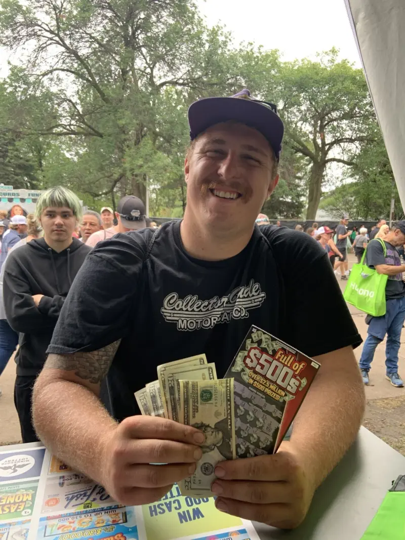 Smiling man in black shirt and cap holds cash and scratch-off ticket at a busy outdoor fair booth.