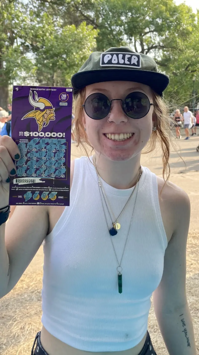 Smiling person in sunglasses, white tank top, and black cap holds a Minnesota Lottery scratch-off ticket outdoors with people and trees behind.