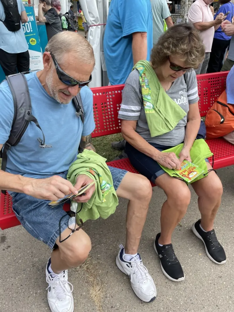 Man and woman in sunglasses sit on a red bench outdoors, scratching off lottery tickets; others are visible in the background.