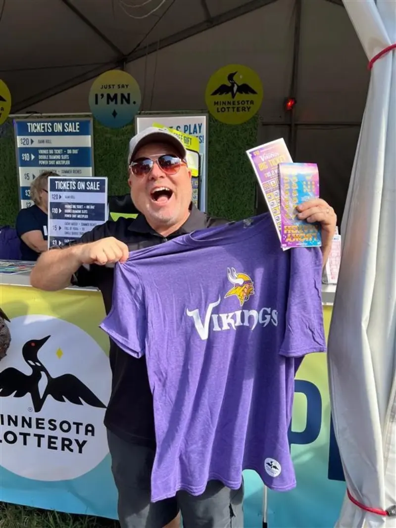 Smiling man in sunglasses and cap holds a Vikings T-shirt and lottery scratch-offs at a Minnesota Lottery booth.