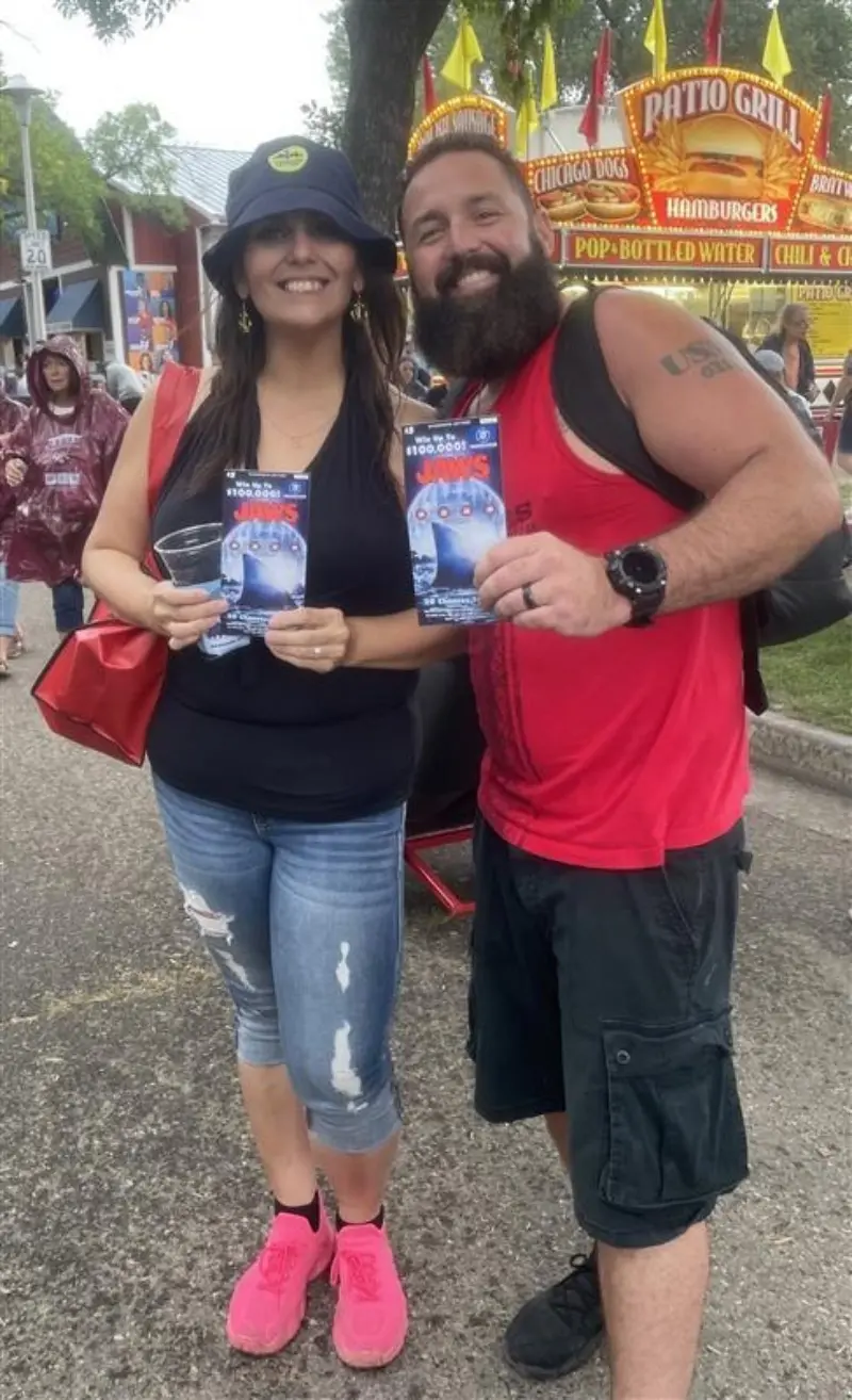Two smiling people hold brochures at a fair, with food stalls and a crowd in the background. Both wear casual, colorful outfits.