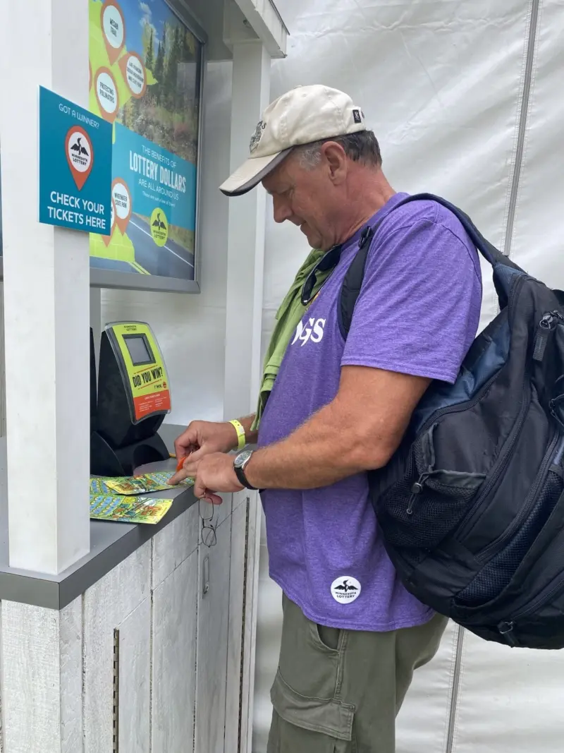 Man in purple shirt and cap scratches a lottery ticket at a kiosk near a display on winnings and ticket checking.
