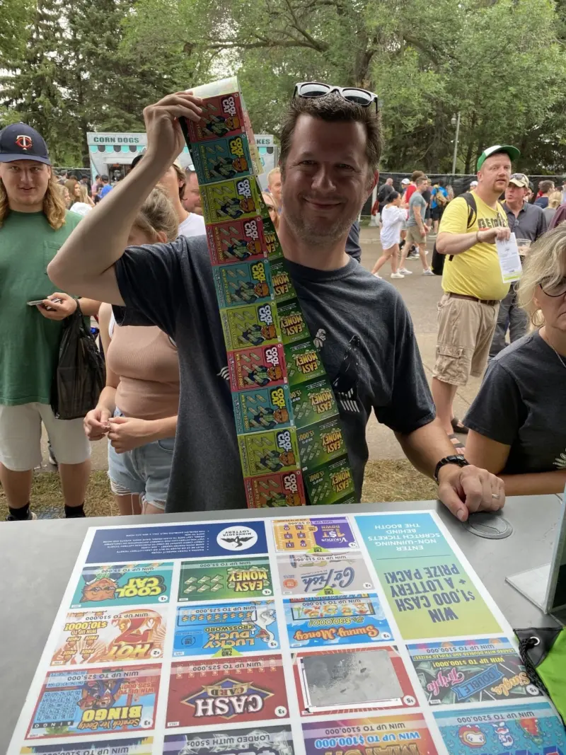 Smiling man holds a long strip of scratch-off tickets at an outdoor fair, with a display of lottery games and attendees behind him.