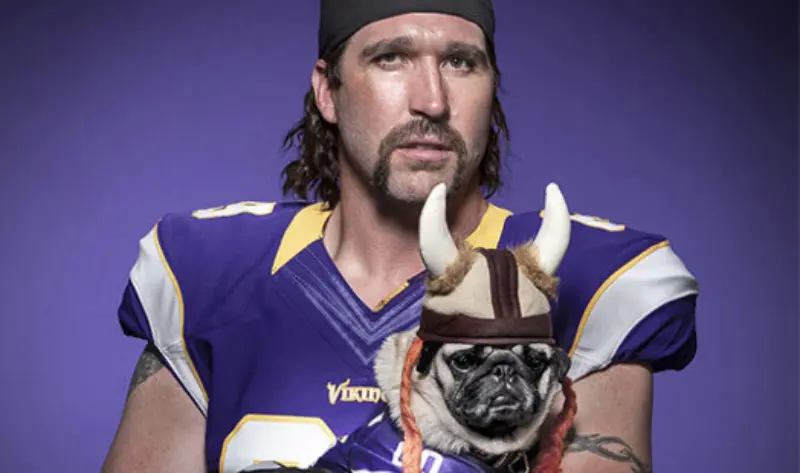 Man in purple football jersey and headband poses with pug in Viking helmet and matching jersey, set against a purple background.