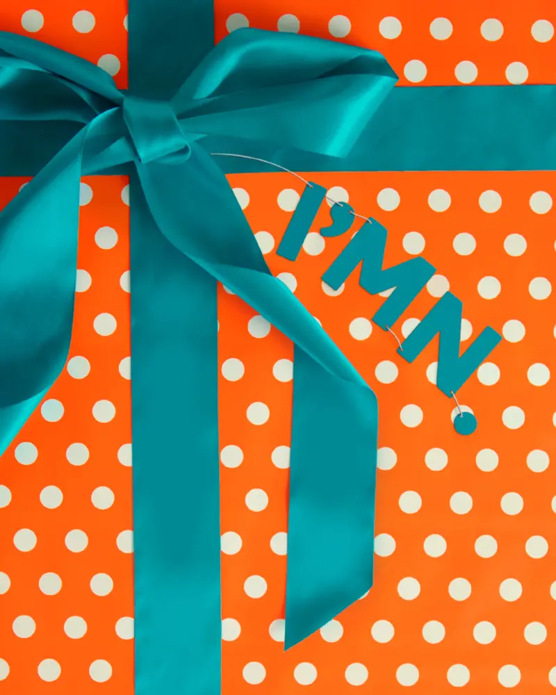 Gift wrapped in orange and white polka dot paper with a blue ribbon; tag reads "P’MN" in blue, referencing the MN State Lottery.
