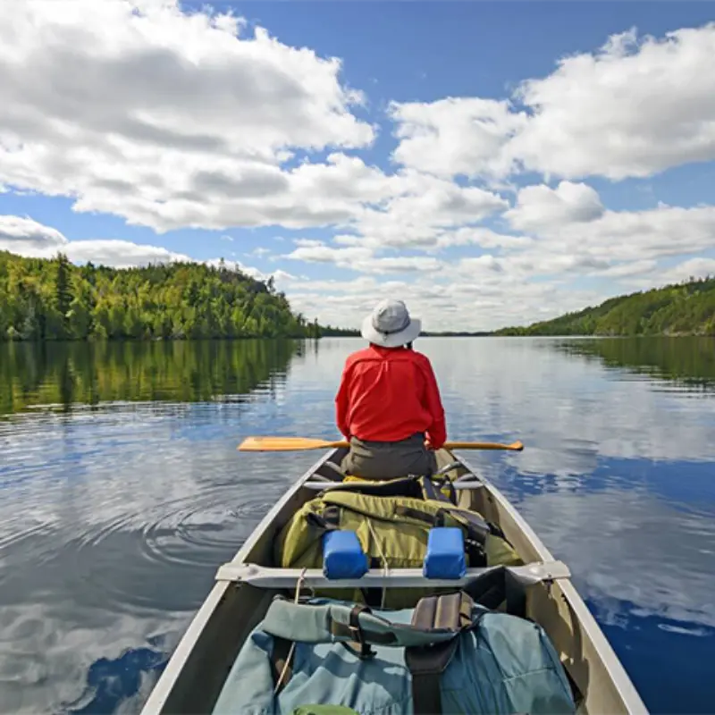 A person in a red shirt and hat paddles a canoe with gear on a calm lake, surrounded by green trees and partly cloudy skies.