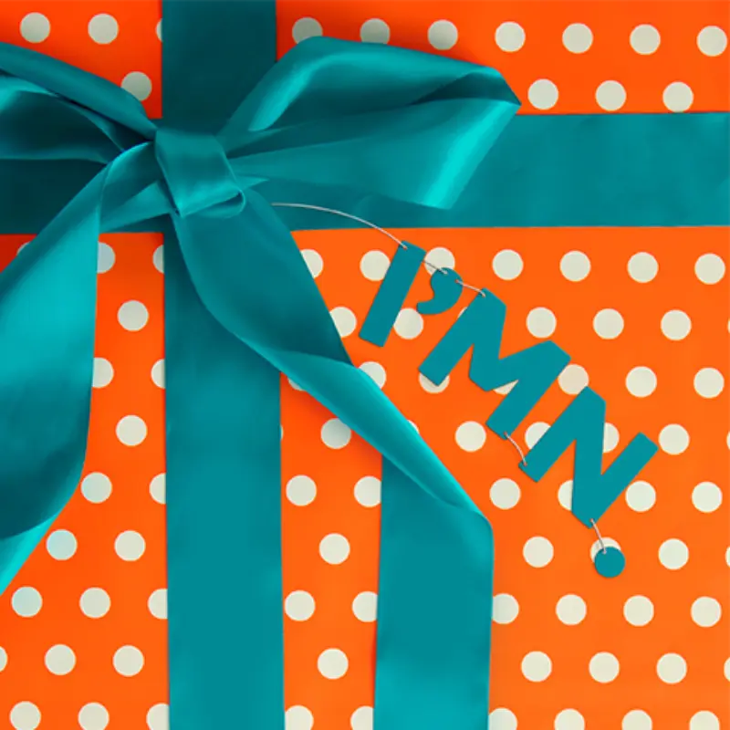 Gift wrapped in orange polka-dot paper with teal ribbon and "IM IN" tag, representing Minnesota Lottery engagement.