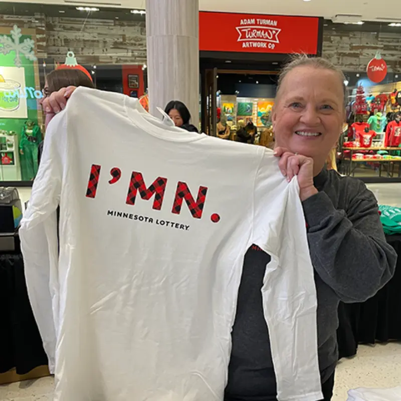 Woman smiling in a mall holds a white long-sleeve shirt with “IMN.” in red plaid and “Minnesota Lottery” below.