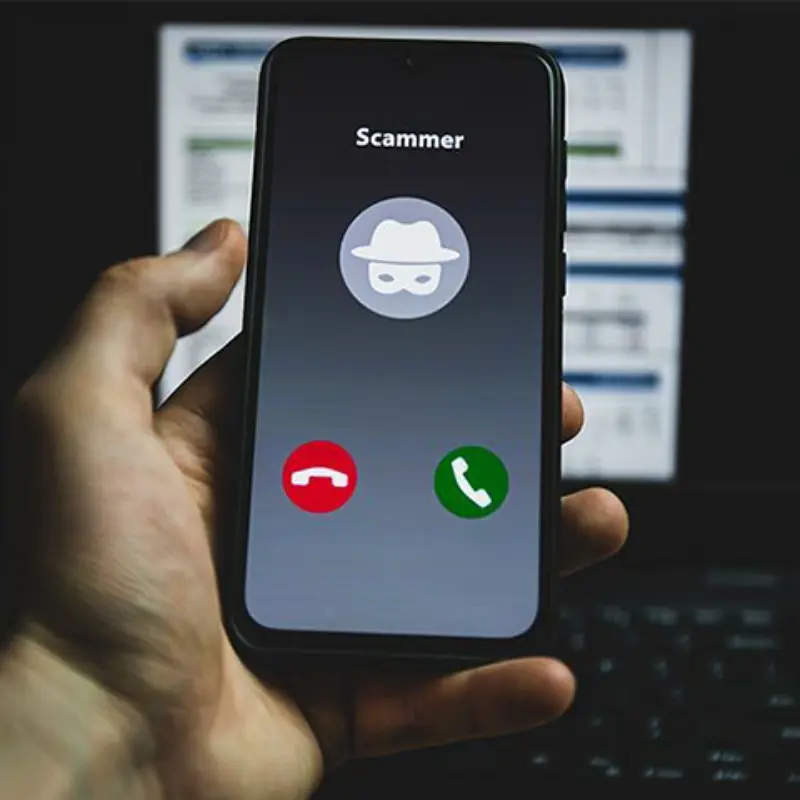 A hand holds a phone showing an incoming call from "Scammer," with red decline, green accept buttons, and a masked caller icon.
