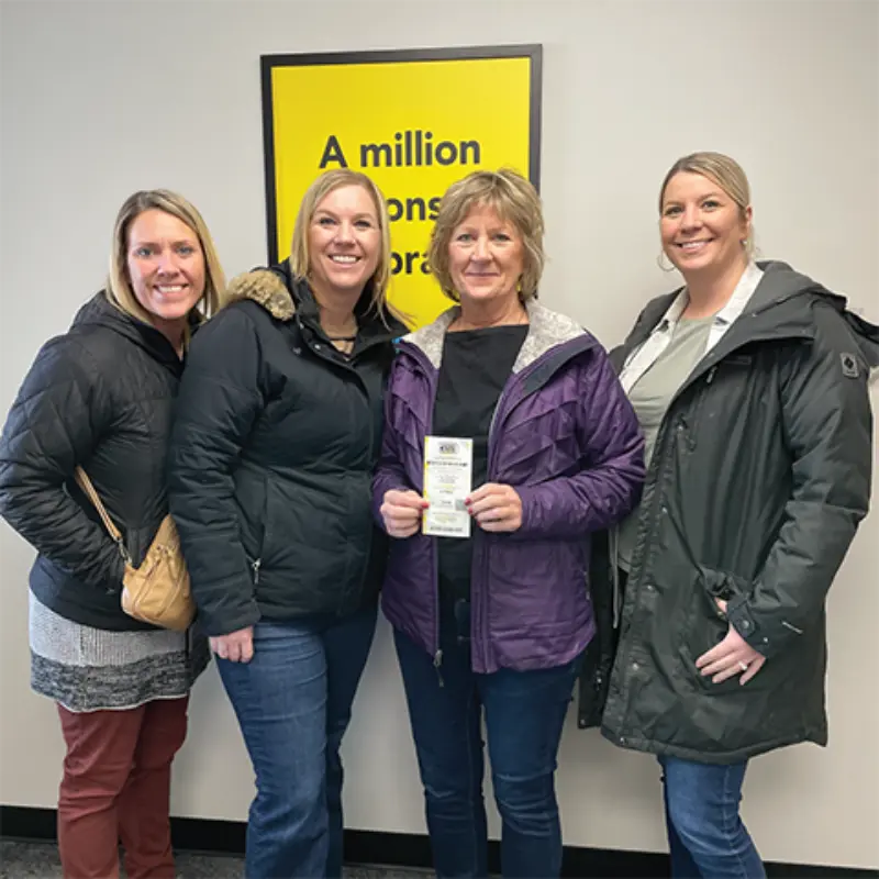 Four smiling women indoors; one holds a lottery ticket. Behind them, a yellow sign reads: "A million reasons to dream.