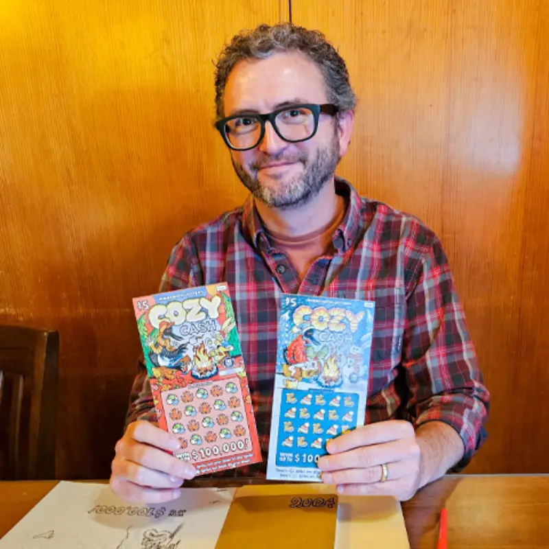 Smiling man in glasses and a red plaid shirt holds up two Cozy Cash lottery scratch-off tickets at a table.