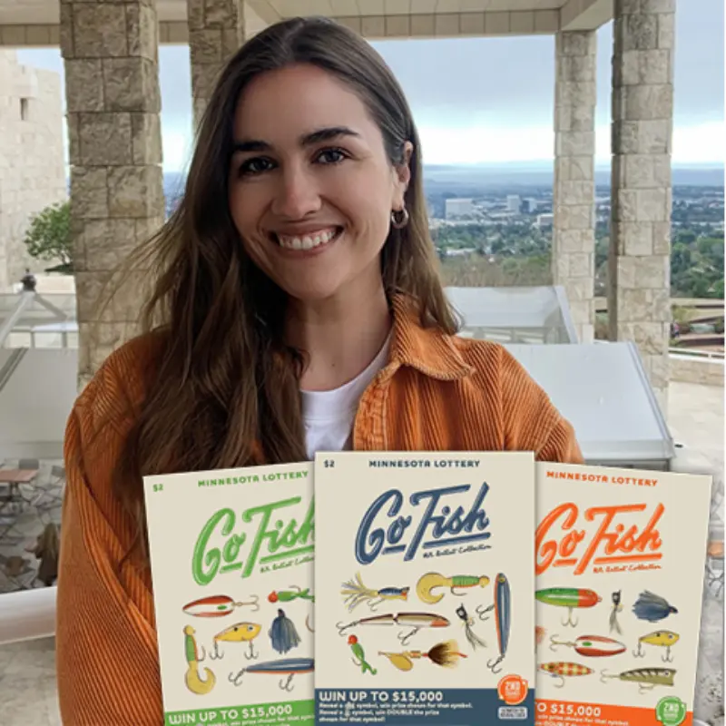 Woman in orange jacket smiles on a balcony, holding three Minnesota Lottery Go Fish tickets with stone architecture in the background.