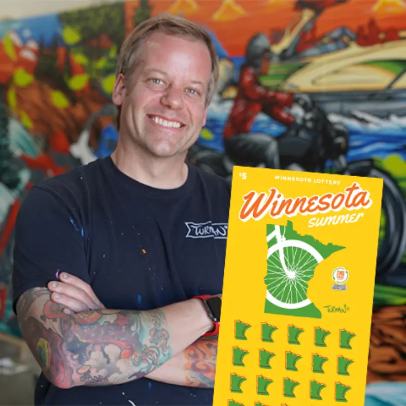 Smiling man stands before a colorful mural, holding a yellow Minnesota Lottery Winnesota Summer scratch-off ticket with green outlines.
