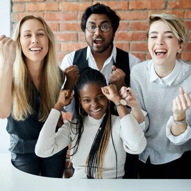 Four people cheer and raise fists in excitement in front of a brick wall, celebrating together with big smiles.