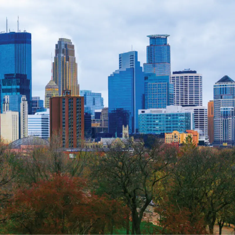 Modern city skyline with tall buildings above autumn trees, under a cloudy sky—representing Minnesota’s vibrant urban landscape.