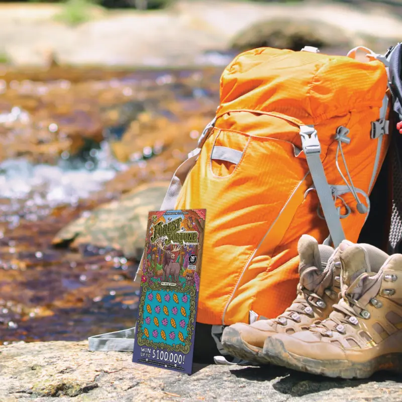 Orange backpack, tan boots, and a Minnesota Lottery scratch-off ticket rest on a rock by a flowing stream outdoors.