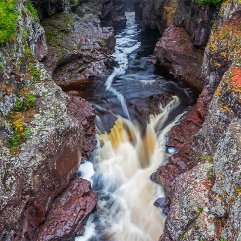 Aerial view of a rocky gorge with rushing dark water, white rapids, mossy cliffs, and green vegetation along the edges.