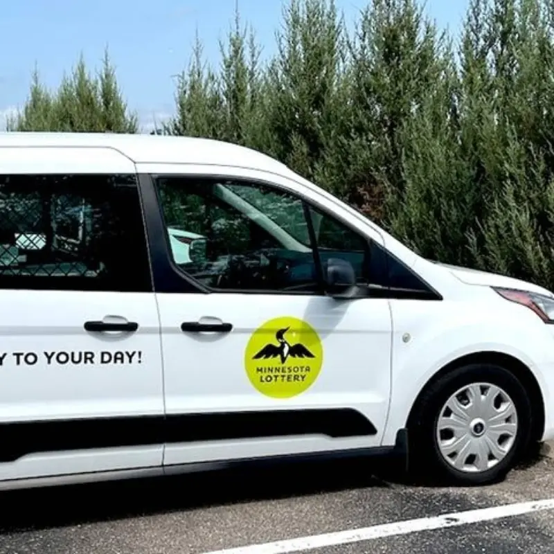 Minnesota Lottery van with logo and loon graphic, parked by trees. Partially visible text on side reads: TO YOUR DAY!.
