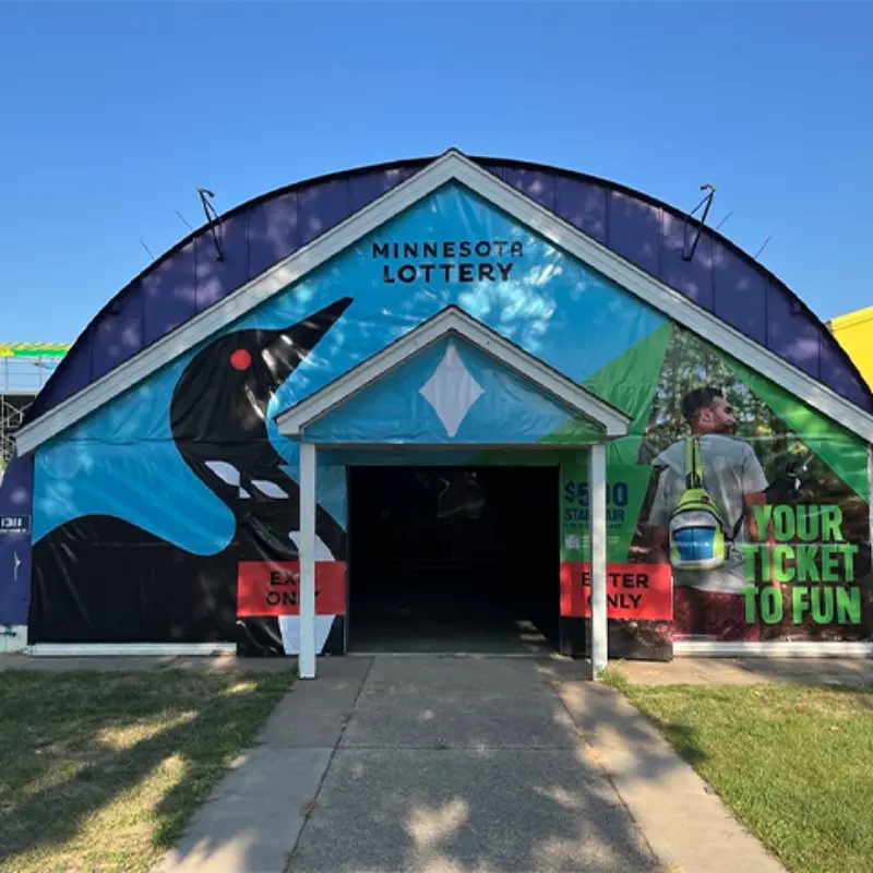 Minnesota Lottery booth with a loon graphic, open entrance, person holding tickets, “Your Ticket to Fun” sign, green grass, and blue sky.