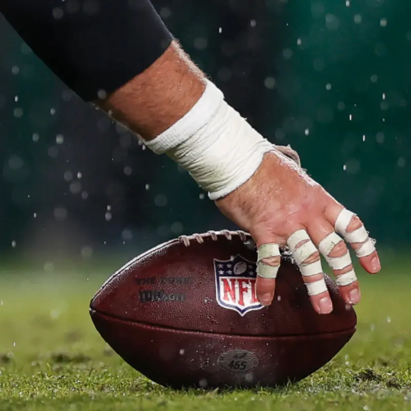 A taped hand readies to snap an NFL football on a wet field, rain droplets in the air—capturing game day excitement.