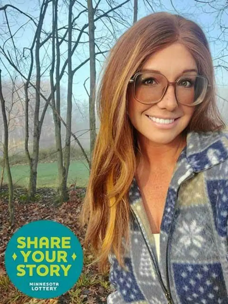 Smiling person with long hair and glasses, in a patterned jacket, stands in misty trees. Share Your Story, Minnesota Lottery emblem is visible.