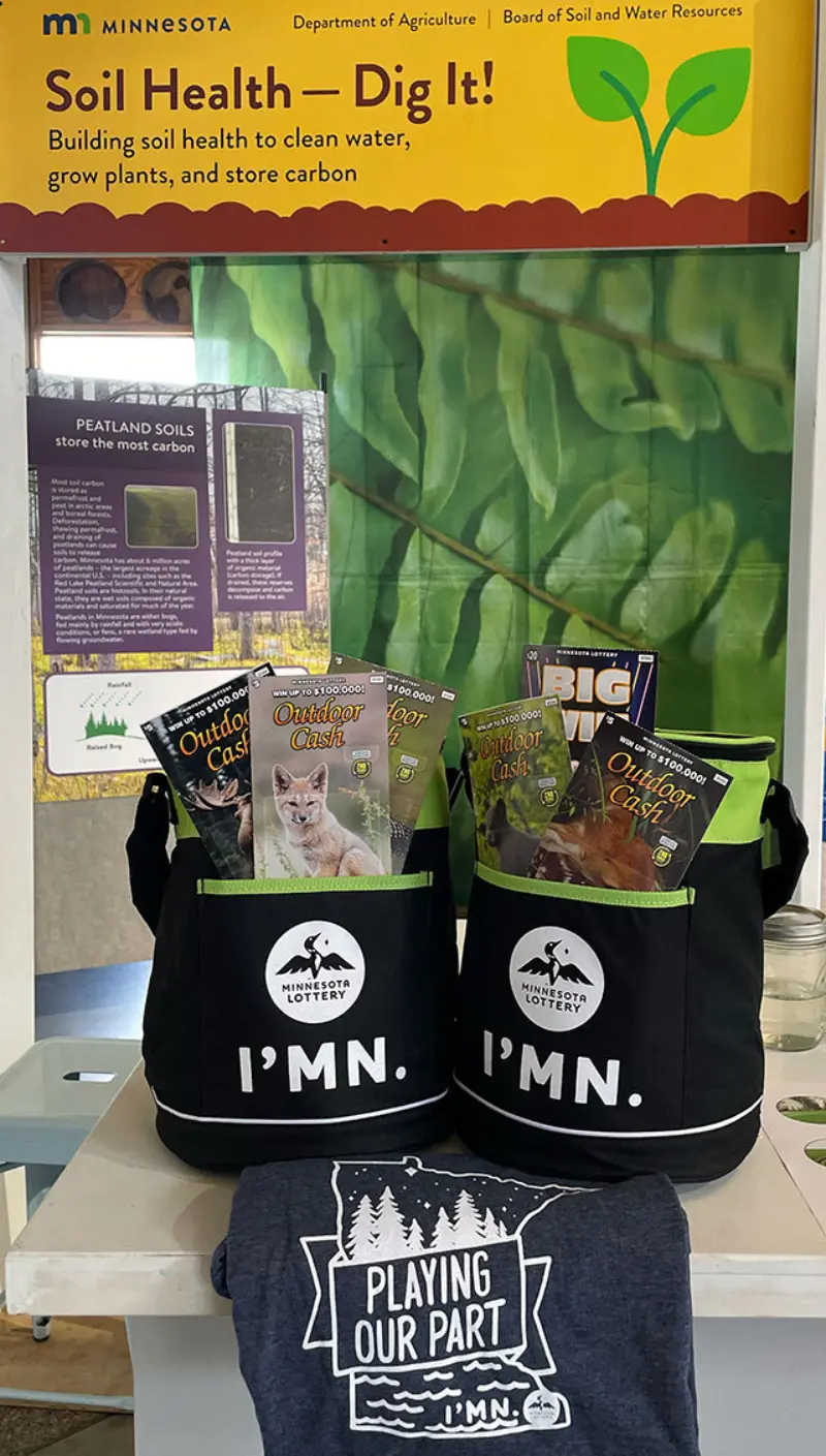 Black IMN. tote bags with outdoor magazines, a Playing Our Part shirt, and soil health sign from MN Dept. of Agriculture on table.