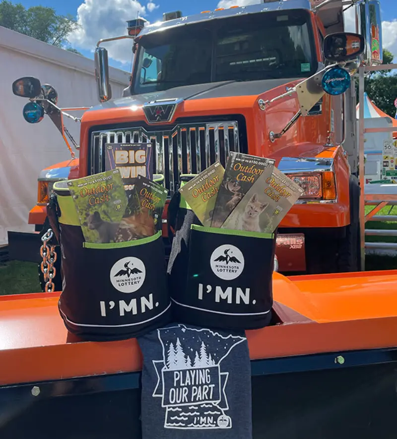 Black IMN bags with Minnesota Lottery tickets sit on an orange truck; a gray Playing Our Part T-shirt is displayed in front.