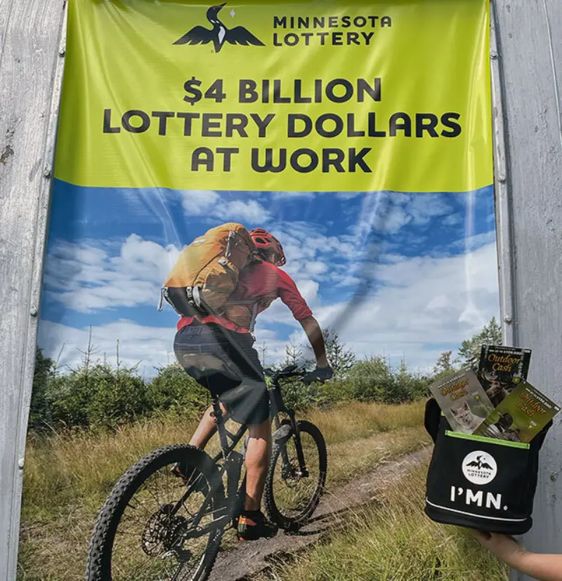 Cyclist passes under “Minnesota Lottery $4 Billion Lottery Dollars At Work” banner; bag with tickets, “I’M IN” can cooler nearby.