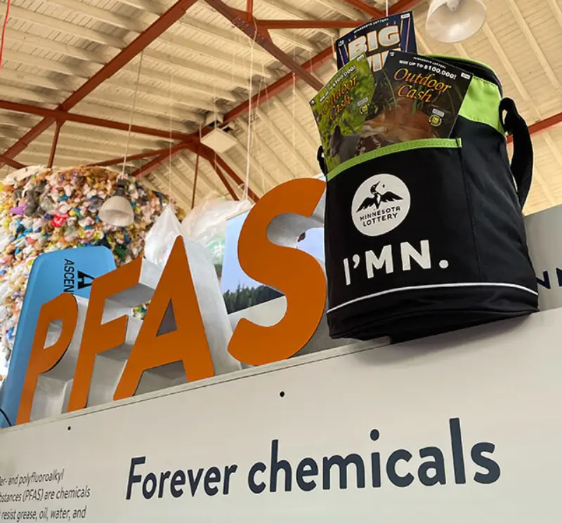 Minnesota Lottery cooler bag with snacks on PFAS Forever Chemicals display at indoor exhibit with colorful decor in the background.