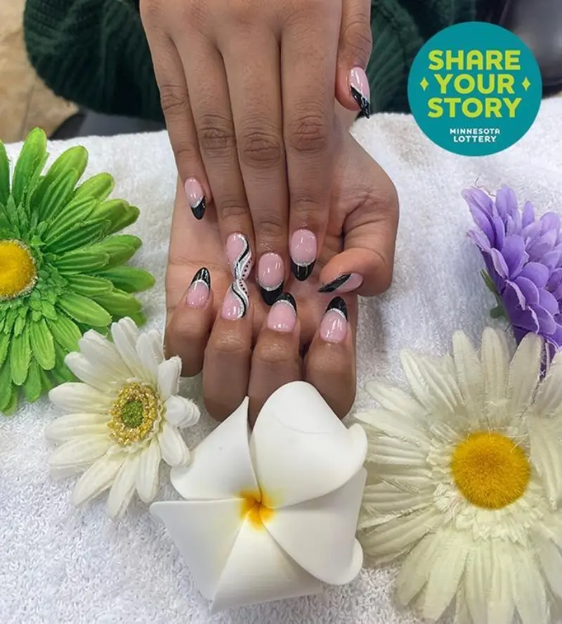 Hands with black and white nail art on a towel, artificial flowers, and a badge reading “SHARE YOUR STORY MINNESOTA LOTTERY.”.