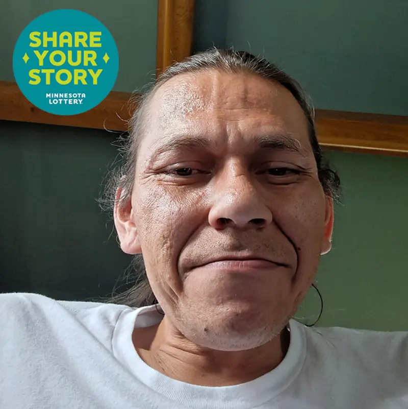 Smiling man with long hair pulled back and white shirt; blue “SHARE YOUR STORY MINNESOTA LOTTERY” circle in corner.