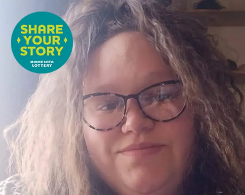 A person with wavy brown hair and glasses smiles at the camera. A blue badge says “Share Your Story Minnesota Lottery.”.